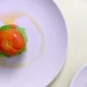 Kuvassa on vaaleanpunaisella lautasella täytetty tomaatti. Kuvan tausta on valkoinen