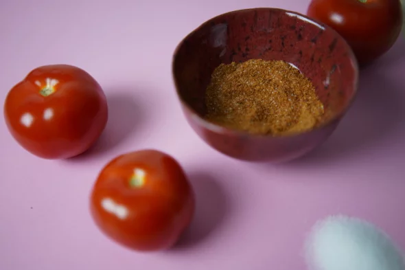 Kuvassa on ruskeassa kipossa tomaattisuolaa. Kipon edessä on kaksi isoa tomaattia. Kuvan tausta on vaaleanpunainen.