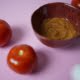 Kuvassa on ruskeassa kipossa tomaattisuolaa. Kipon edessä on kaksi isoa tomaattia. Kuvan tausta on vaaleanpunainen.